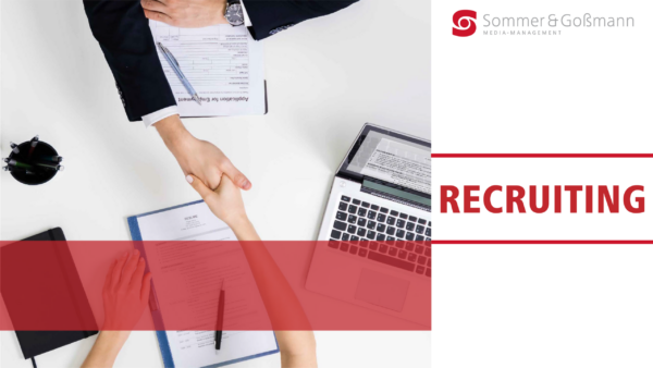 Zwei Personen, die sich über einem Schreibtisch die Hand geben, umgeben von Dokumenten, einem Laptop und Schreibmaterial